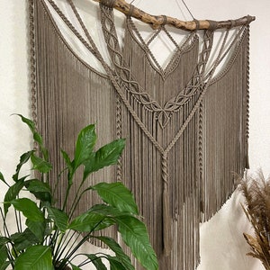 May include: A large macrame wall hanging with a natural wood branch at the top. The macrame is made with brown cord and has a fringe at the bottom. The wall hanging is hanging on a white wall.