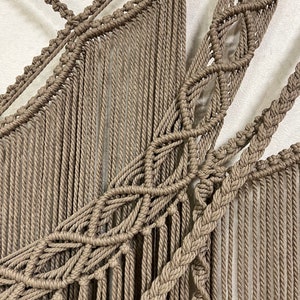May include: A beige macrame wall hanging with a geometric pattern. The hanging is made of thick rope and has a fringe at the bottom.