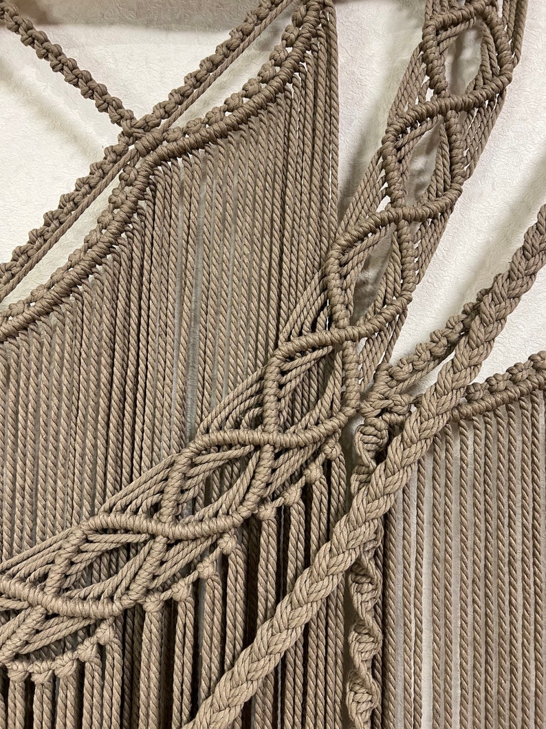 May include: A beige macrame wall hanging with a geometric pattern. The hanging is made of thick rope and has a fringe at the bottom.