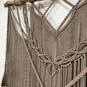 May include: A brown macrame wall hanging with a wooden branch at the top. The macrame is made with a variety of knots and patterns, and the fringe is long and flowing.