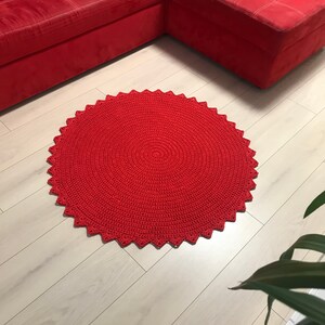 May include: A red crocheted round rug with a scalloped edge. The rug is on a light wood floor.