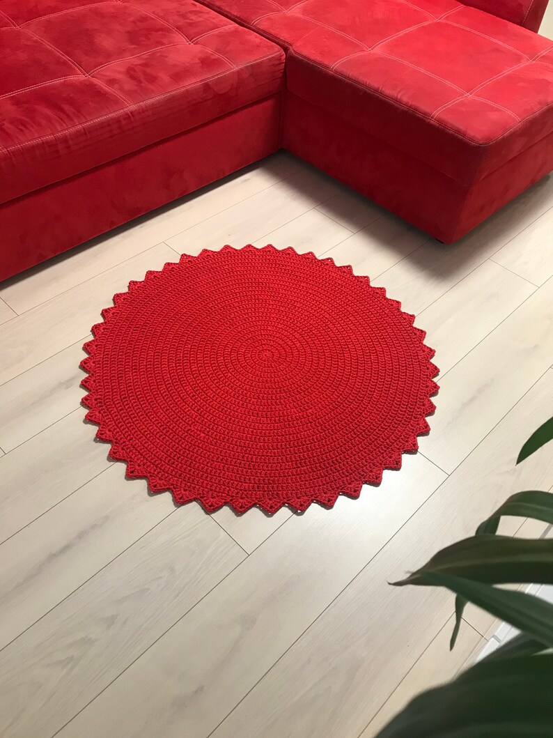 May include: A red crocheted round rug with a scalloped edge. The rug is on a light wood floor.