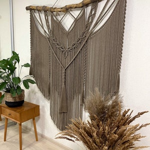 May include: A large, brown macrame wall hanging with a wooden branch at the top. The hanging features a geometric design with tassels at the bottom.