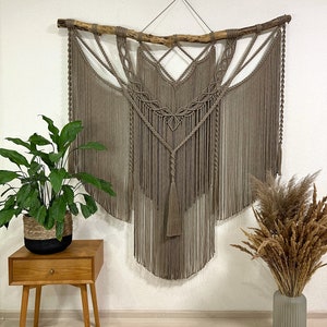 May include: A large macrame wall hanging with a natural brown color. The hanging is made with a thick cord and features a geometric design. The hanging is attached to a natural branch.