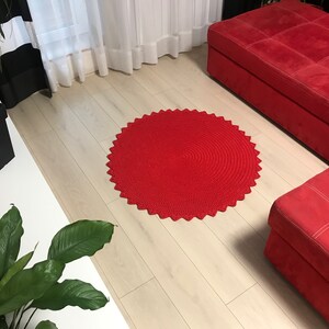 May include: A red crocheted round rug with a scalloped edge. The rug is on a light wood floor in a living room setting.