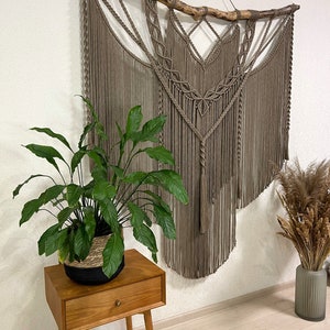 May include: A large macrame wall hanging with a natural wood branch at the top. The macrame is made with thick brown cord and features a geometric pattern. The wall hanging is hanging on a white wall and there is a potted plant and a vase with dried flowers in the foreground.