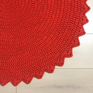 May include: A close-up of a red crocheted rug with a scalloped edge. The rug is made of thick yarn and has a textured surface.