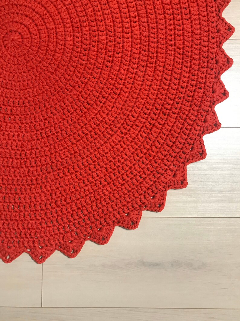 May include: A close-up of a red crocheted rug with a scalloped edge. The rug is made of thick yarn and has a textured surface.