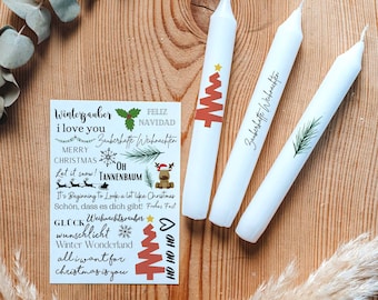 Op de afbeelding: Drie witte taps toelopende kaarsen met kerstontwerpen en een stickervel met vakantie zinnen en illustraties. De kaarsen hebben een rode kerstboom, een dennentak en tekst. Het stickervel bevat zinnen als "Vrolijk Kerstfeest" en "Ik hou van jou."