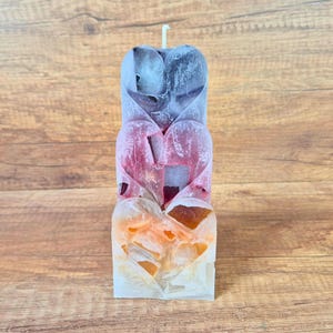 Puede incluir: Una vela de pilar rectangular con un diseño de corazones apilados. La vela cambia de color desde un morado oscuro en la parte superior, pasando por un rosa vibrante en el centro, hasta un naranja cálido y blanco en la base, con una mecha.