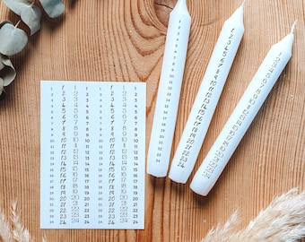 May include: Three white candles with black numbers printed on them. The candles are numbered 1 through 24. The candles are on a wooden surface with a sprig of eucalyptus and a dried grass plume.