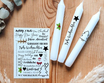 May include: White taper candles with festive designs, including a star, holly, and Christmas tree. A card with holiday greetings in German and English is also present. The arrangement is set on a wooden surface, evoking a seasonal theme.