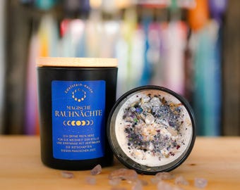 May include: A black candle in a jar with a bamboo lid and a blue label featuring the text Magische Rauhnächte and moon phases. The open candle reveals white wax topped with various small crystals, dried herbs, and dark glitter, with loose stones nearby.