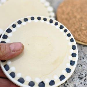 Könnte beinhalten: Ein weißer Keramikuntersetzer mit einem schwarzen und weißen Tupfenrand. Der Untersetzer ist rund und hat eine glatte Oberfläche.