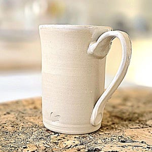 Könnte beinhalten: Eine weiße Keramiktasse mit einem abgerundeten Henkel. Die Tasse hat eine dezente gesprenkelte Textur und ein kleines Blattdesign an der Seite.