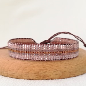 May include: A pink and copper beaded bracelet with a brown cord closure. The bracelet is on a wooden surface.