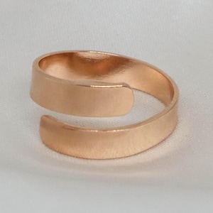 May include: A rose gold-colored, adjustable ring. The ring features a simple, minimalist design with a flat band that wraps around the finger. The ring is made of a shiny metal and is set against a white background.