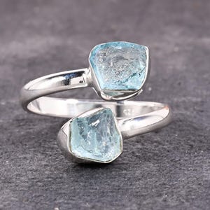 May include: A silver ring with two rough-cut, pale blue aquamarine gemstones. The ring's band wraps around the finger, with one stone at the top and the other at the bottom. The stones have a natural, unpolished look, set against a dark grey background.