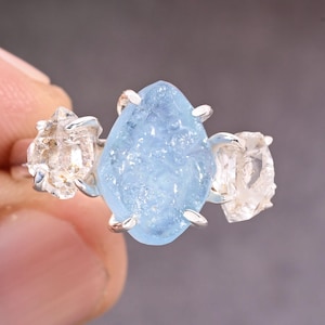 Puede incluir: Un anillo de plata con una gema grande, azul claro y en bruto como pieza central, flanqueada por dos piedras más pequeñas, transparentes y en bruto. El anillo se sostiene sobre un fondo borroso.