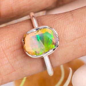 Op de afbeelding: Een zilveren ring met een ovale opaal edelsteen. De opaal toont een levendig kleurenpalet, waaronder groen, geel en oranje. De ring heeft een eenvoudige, gepolijste zilveren band. De edelsteen is gezet in een bezel setting.
