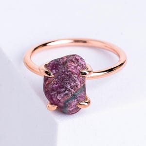 Könnte beinhalten: Ein roségoldener Ring mit einem rohen Rubin-Edelstein. Der Rubin hat eine tiefe Magenta-Farbe mit sichtbarer Textur und natürlichen Variationen. Der Ring hat ein schlichtes, dünnes Design, das die einzigartige Form des Edelsteins hervorhebt.