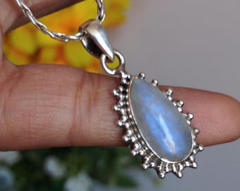 May include: A silver necklace with a teardrop-shaped pendant. The pendant features a large, oval, blue gemstone with a silver border. The chain is a thin, twisted design. The background is blurred, with hints of yellow and green.