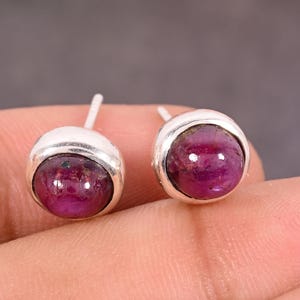 May include: A pair of silver stud earrings, each with a round, polished, deep red gemstone. The gemstones are set in a simple bezel setting. The earrings are shown against a neutral background.
