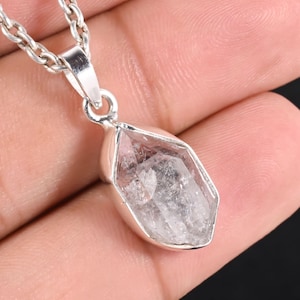 Pu&ograve; includere: Una collana con pendente in cristallo di quarzo trasparente con una catena d'argento. Il cristallo di quarzo &egrave; sfaccettato e incastonato in una lunetta d'argento. La collana &egrave; tenuta in mano su sfondo scuro.