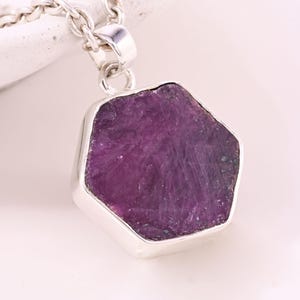 May include: A silver necklace with a hexagonal pendant showcasing a raw, deep purple gemstone. The pendant is set in a silver bezel and suspended from a delicate silver chain. The gemstone has a textured surface.