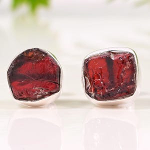 Pode incluir: Um par de brincos de press&atilde;o com pedras de granada vermelha escura bruta, emolduradas em prata. As pedras preciosas t&ecirc;m uma textura natural &aacute;spera. Exibido em um fundo branco com folhagem verde.