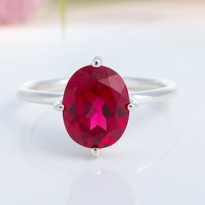 Könnte beinhalten: Ein silberner Ring mit einem großen, oval geschliffenen Rubin. Der leuchtend rote Stein ist der Mittelpunkt, gefasst in einem schlichten, eleganten Band. Das Design des Rings umfasst kleine dekorative Akzente in der Nähe des Edelsteins.