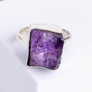 May include: A silver ring with a large, rectangular, raw amethyst gemstone. The amethyst is a deep purple colour with a textured surface. The ring is set on a white surface.