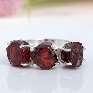 May include: A silver ring featuring three raw, deep red garnet stones. The gemstones are irregularly shaped and set in individual prongs, creating a unique and naturalistic design. The ring is displayed on a white surface.