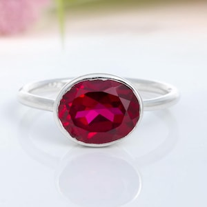 May include: A silver ring featuring a large, oval-cut, deep red gemstone. The gemstone is the central element, set in a simple, polished band. The ring is presented on a white surface, with blurred floral accents in the background.