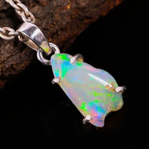 Könnte beinhalten: Eine silberne Halskette mit einem Anhänger aus einem rohen, schillernden Opal-Edelstein. Der Opal zeigt grüne, blaue und rosa Farbtöne. Der Anhänger wird von silbernen Zinken gehalten.