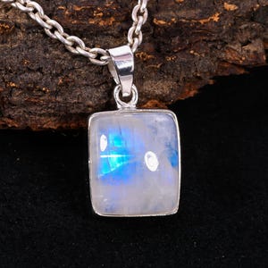Pode incluir: Um colar de prata com um pingente quadrado de pedra da lua iridescente. O pingente exibe uma mistura de tons azuis e brancos. A corrente é de estilo clássico e o pingente é fixado em uma moldura de prata.