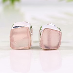 Puede incluir: Un par de pendientes cuadrados con montura plateada y una piedra translúcida de color rosa claro. Los pendientes se exhiben sobre una superficie blanca con un fondo verde borroso.