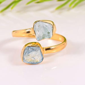 May include: A gold ring with two light blue aquamarine gemstones. The ring has a bypass design, with the stones set in a bezel setting. The aquamarine stones have a rough, natural appearance.