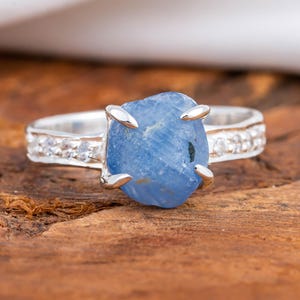 May include: A silver ring featuring a large, rough-cut, light blue gemstone as the centerpiece. The band is detailed with small, clear stones. The ring is set against a wooden surface, with a white fabric in the background.