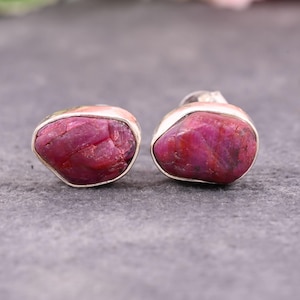 Peut inclure: Une paire de boucles d'oreilles à clous avec des pierres précieuses de rubis brutes et naturelles, serties dans un sertissage en argent. Les rubis sont d'un rouge profond et ont une surface rugueuse et texturée.