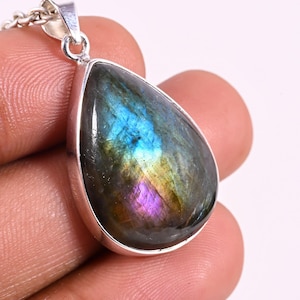 Puede incluir: Un colgante de labradorita en forma de lágrima engastado en un bisel de plata pulida. La piedra preciosa muestra destellos iridiscentes de azul, oro y púrpura sobre una base gris oscuro. El colgante está unido a una anilla de plata.