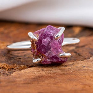 May include: A silver ring featuring a raw, uncut ruby gemstone. The ruby is a deep pinkish-red color and is held in place by four silver prongs. The ring band is a simple, polished silver.