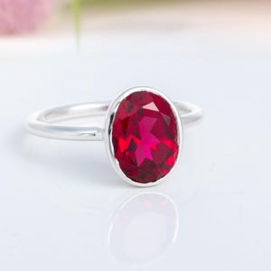 May include: A silver ring featuring a large, oval-cut, deep red gemstone. The gemstone is set in a bezel setting, and the band is smooth and polished. The ring is displayed on a white surface, with a blurred floral background.