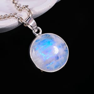 May include: A round, iridescent moonstone pendant necklace. The moonstone has a blue and white color scheme. The pendant is set in a silver-colored metal and hangs from a matching chain. The necklace is displayed against a dark background.