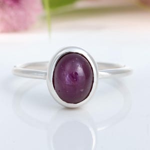 Op de afbeelding: Een zilveren ring met een grote, ovale, dieppaarse edelsteen. De edelsteen is gezet in een bezel setting en de band is dun en eenvoudig. De ring wordt gepresenteerd op een wit oppervlak, met een vage bloemenachtergrond.