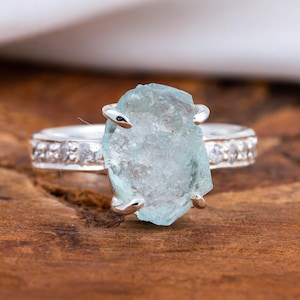 Pode incluir: Um anel de prata com uma grande pedra preciosa de água-marinha azul clara. A banda é adornada com pequenas pedras transparentes. A pedra preciosa é mantida por quatro garras de prata. O anel está sobre uma superfície de madeira.