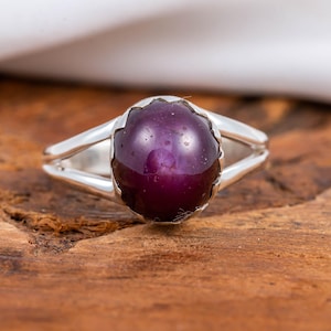 Peut inclure: Une bague en argent ornée d'une grosse pierre précieuse ovale, violet foncé. La pierre est sertie dans une lunette festonnée et présente une surface polie et réfléchissante. L'anneau de la bague se divise en deux lignes parallèles.