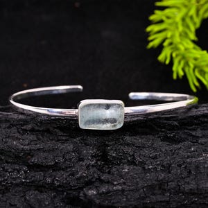 May include: A silver cuff bracelet featuring a rectangular, light blue gemstone. The bracelet has a simple, open design with a polished finish. The gemstone is set in a bezel setting, and the bracelet is displayed on a dark, textured surface.