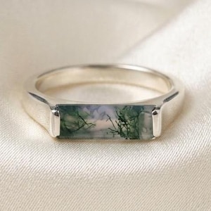 Puede incluir: Un anillo de plata con una piedra de ágata musgosa rectangular montada horizontalmente. La piedra muestra tonos blancos y grises translúcidos con inclusiones de color verde oscuro similares a ramas. El aro es liso, pulido y sencillo.