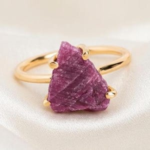 May include: A gold ring featuring a raw, triangular-shaped deep pink gemstone set in a prong setting. The stone has a textured, crystalline surface with visible natural facets, while the band is smooth, polished, and circular in design.